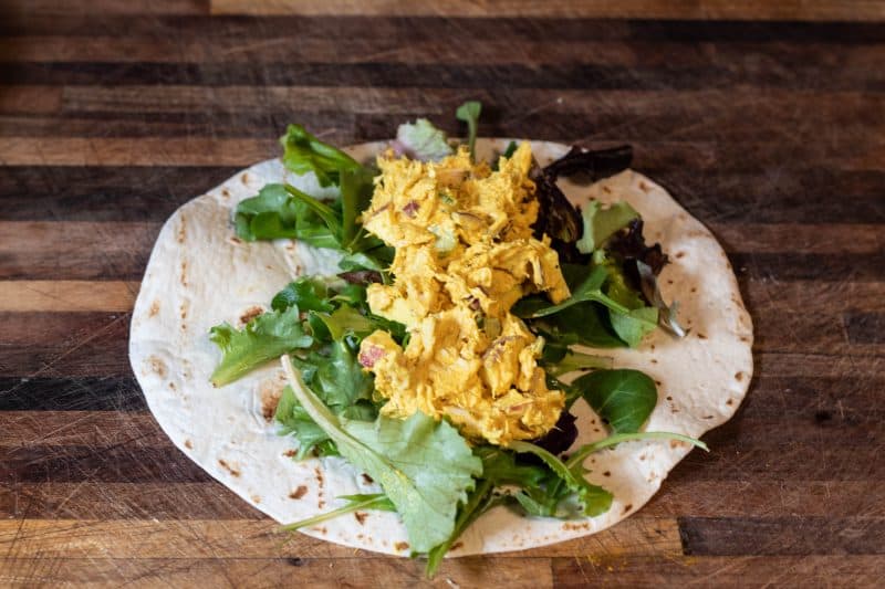 Tortilla with spring mix and curry chicken salad on a wood cutting board.