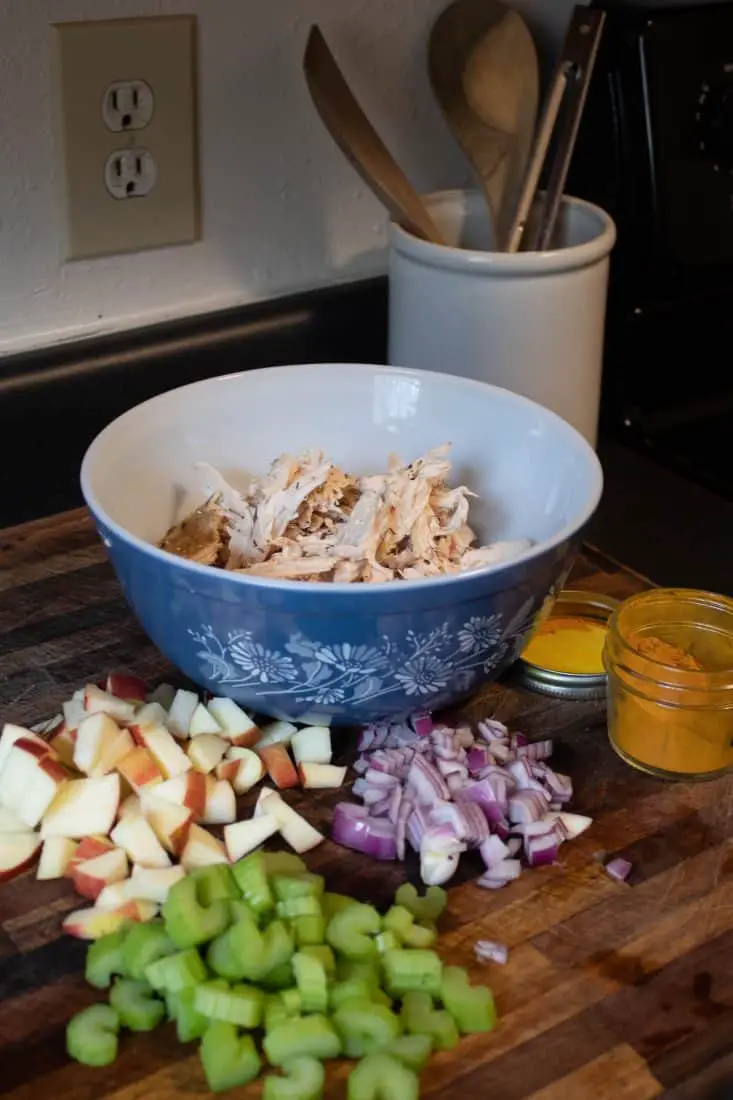 Image of all the ingredients to curry chicken salad on a cutting board. Diced red onion, apples, and celery with shredded chicken in a bow a container of powered curry.