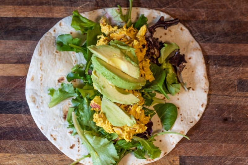 Tortilla with spring mix, curry chicken salad, and sliced avocado on a wood cutting board.